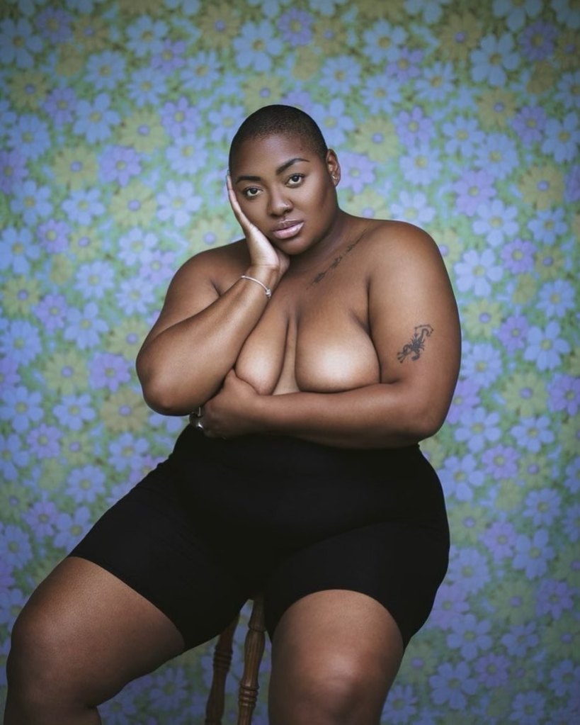 Image shot my Alex Cameron. Nyome is looking directly into camera. One hand rests on her cheek, the other arm and hand covers her breasts. She is wearing black cycle shorts, is sitting on a stool, and is sat in front of a green and purple floral backdrop