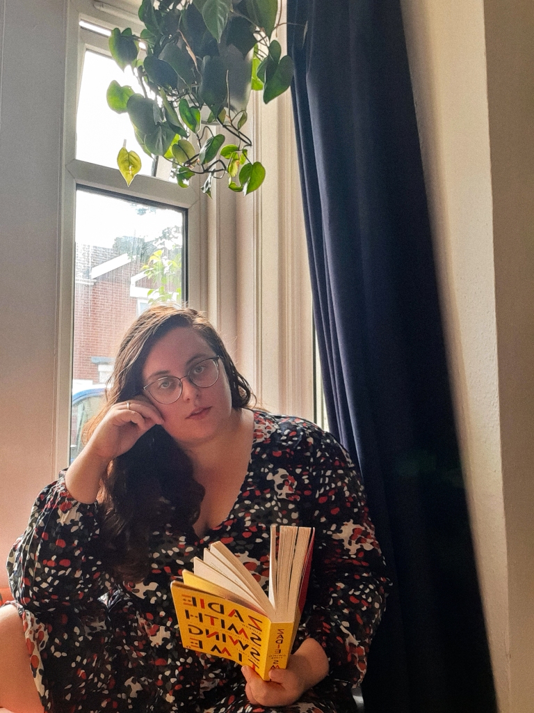 Rosie is sitting in a white window nook, next to a navy curtain. She has dark hair, glasses, and is looking directly up at the camera. One hand is resting on her cheek, the other is holding open a yellow book - it is Zadie Smith's Swing Time.