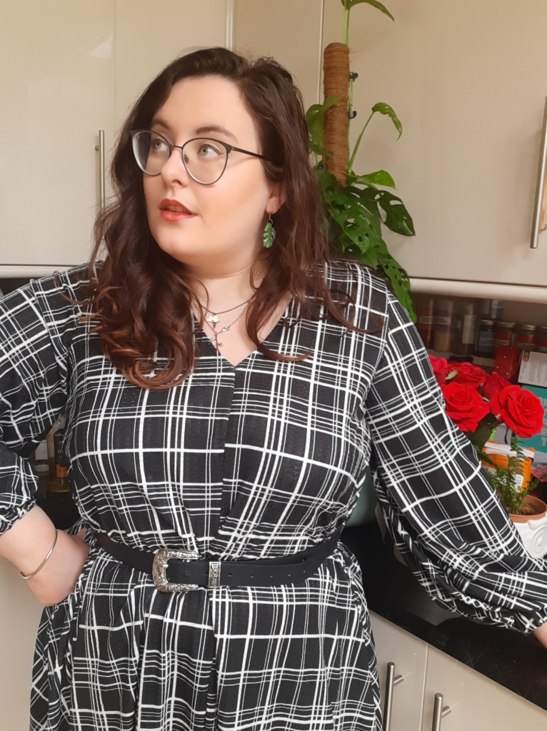 Rosie is looking to the right and has her hand on her hip. She is wearing a long sleeve, black and white check midi dress which has a v-neck. She is also wearing a black Western belt.
