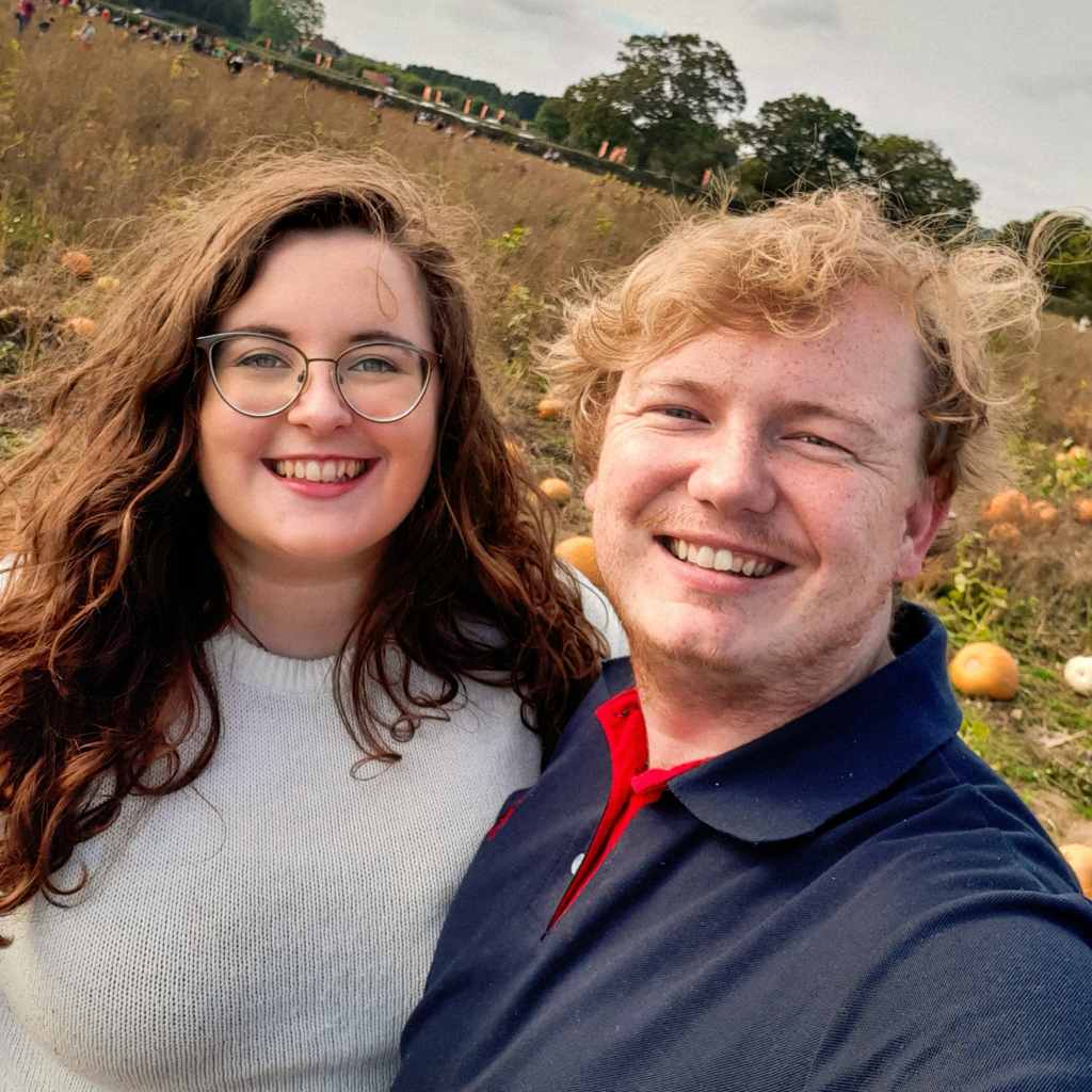A picture of Rosie and Rory smiling in a pumpkin field. They are both Caucasian. Rosie has brown wavy hair, glasses, and is wearing a white jumper. Rory has dark blonde, short wavy hair, ginger stubble, and is wearing a navy polo shirt with a red interior.