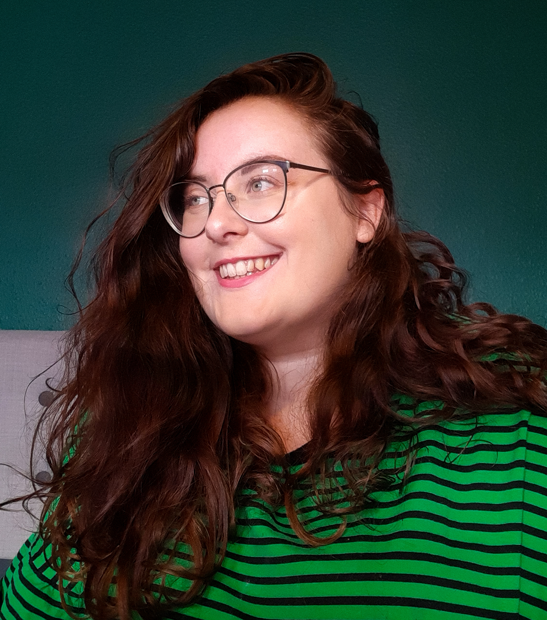A picture of Rosie looking to the left. She has brown wavy hair and is Caucasian. She is wearing large, thin black glasses, and a green and black striped top.