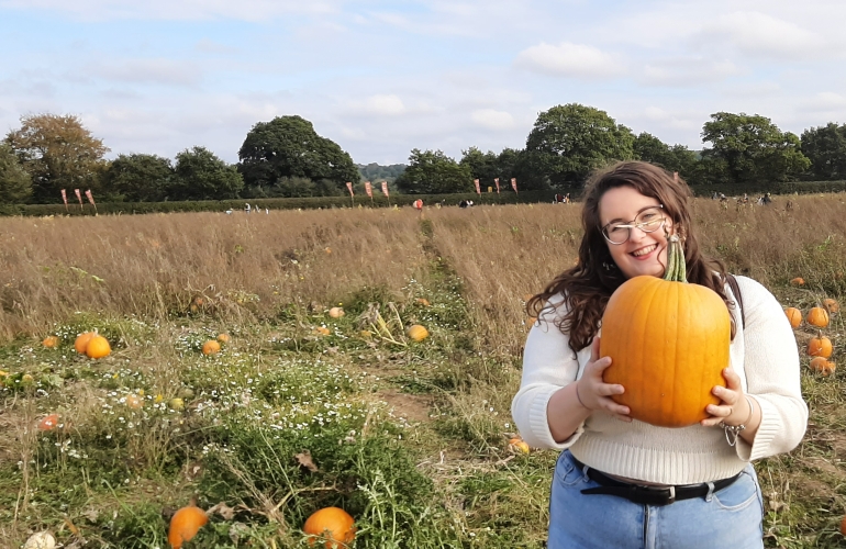 An image of a green and brown field. Rosie is in the forefront holding a pumpkin.