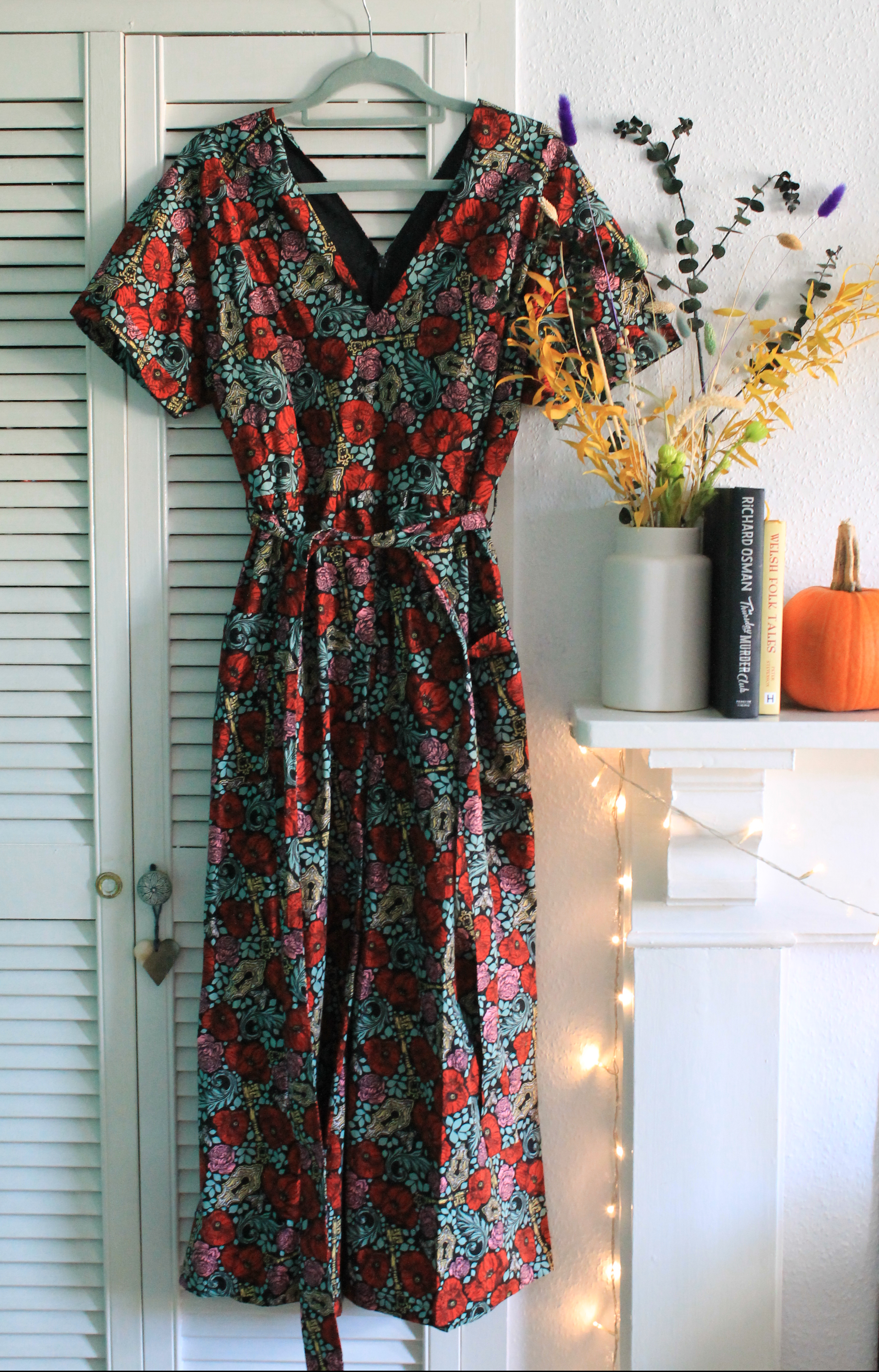 A jumpsuit hanging on a white cupboard. The jumpsuit is printed with red, green, and pink flowers. It is next to a white vase full of dried flowers.