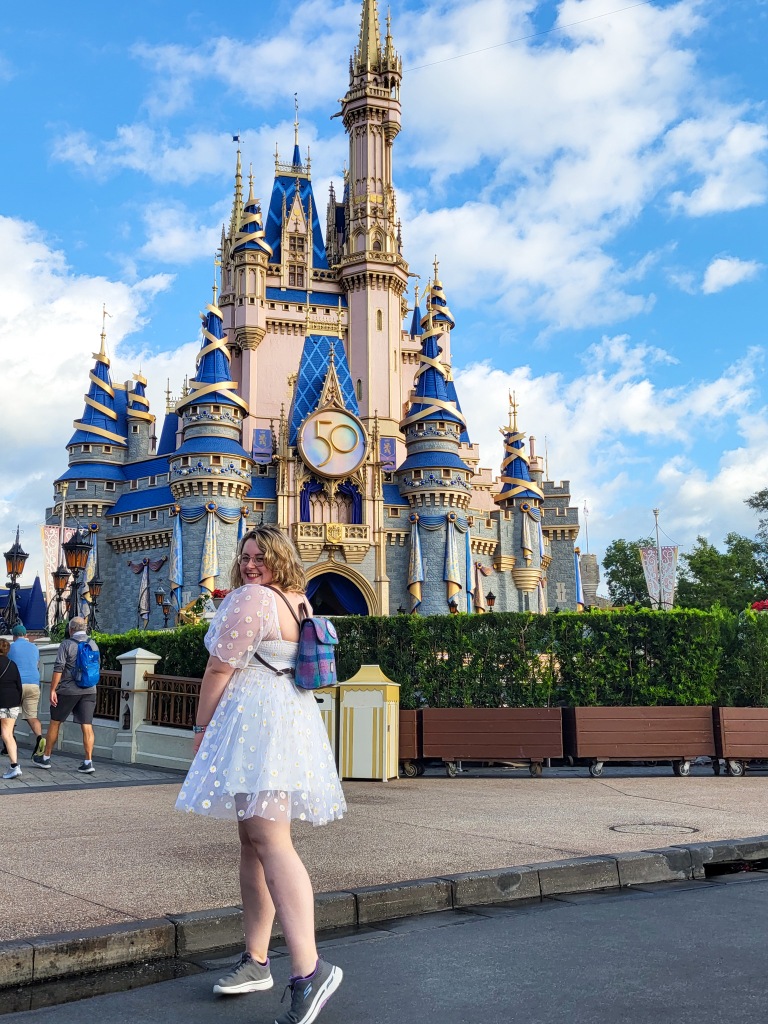 An image of Rosie standing in front of Cinderella Castle. Rosie is a white, chubby woman, with a blonde wavy bob and glasses. She is looking over her left shoulder into the camera. She is wearing a white minidress and grey trainers. The castle is pale pink with blue accents and the sky behind it is blue with white clouds. 