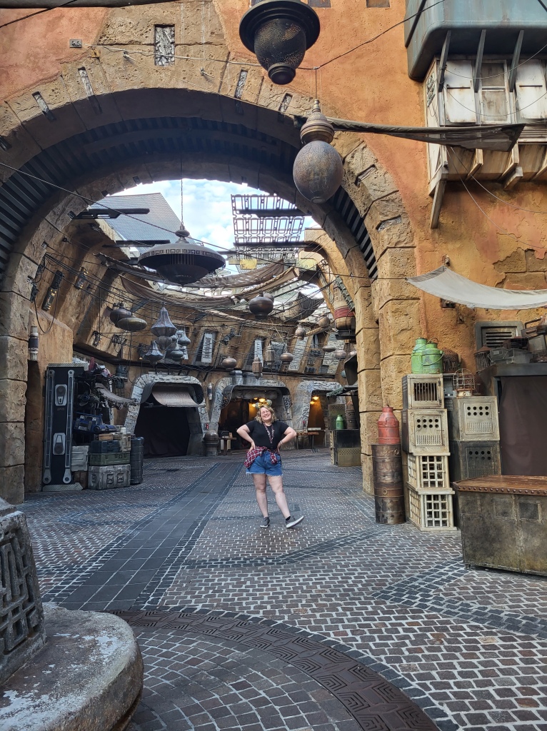 An image of Rosie stood underneath an archway in Batuu, a Star Wars themed land. The ground is paved with grey brick and the walls are yellow sandstone. Rosie is wearing denim shorts, a black tee, and has a red check shirt around her waist. 