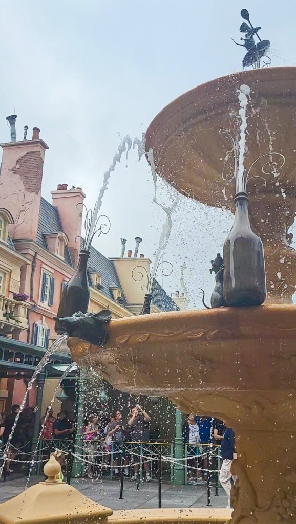 An image of a sandstone fountain spraying water from wine bottles. The bottles are being held up by carved rats. Behind the fountain is a façade of a French town. 