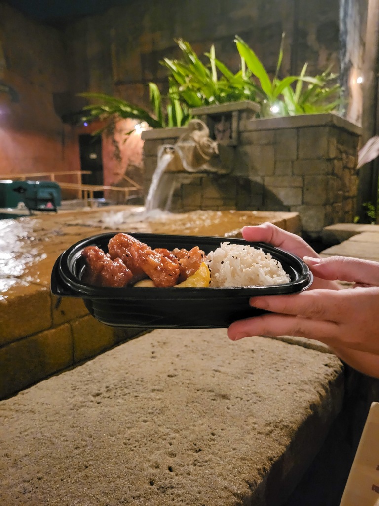 An image of two white hands holding a black bowl of chicken, vegetables and rice. Behind the bowl is a fountain and greenery. 