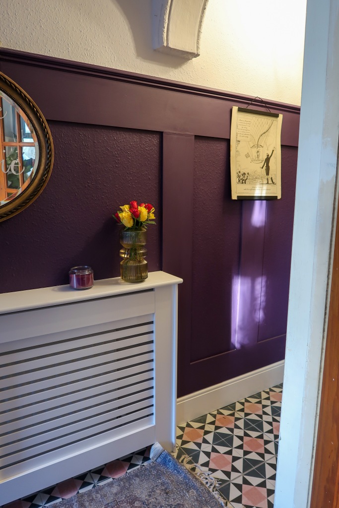 An image of our hallway taken through a doorway. The tiles are pink, blue and white; the walls are panelled and purple. There is a white radiator cover, and a Victorian print on the wall. 