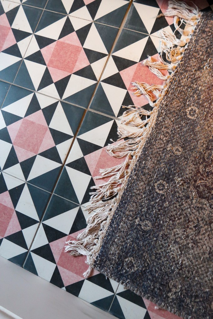 Moroccan style porcelain tiles on the floor. They are pink, black,navy and white in a triangle pattern. On top of the floor is a purple Persian style rug.