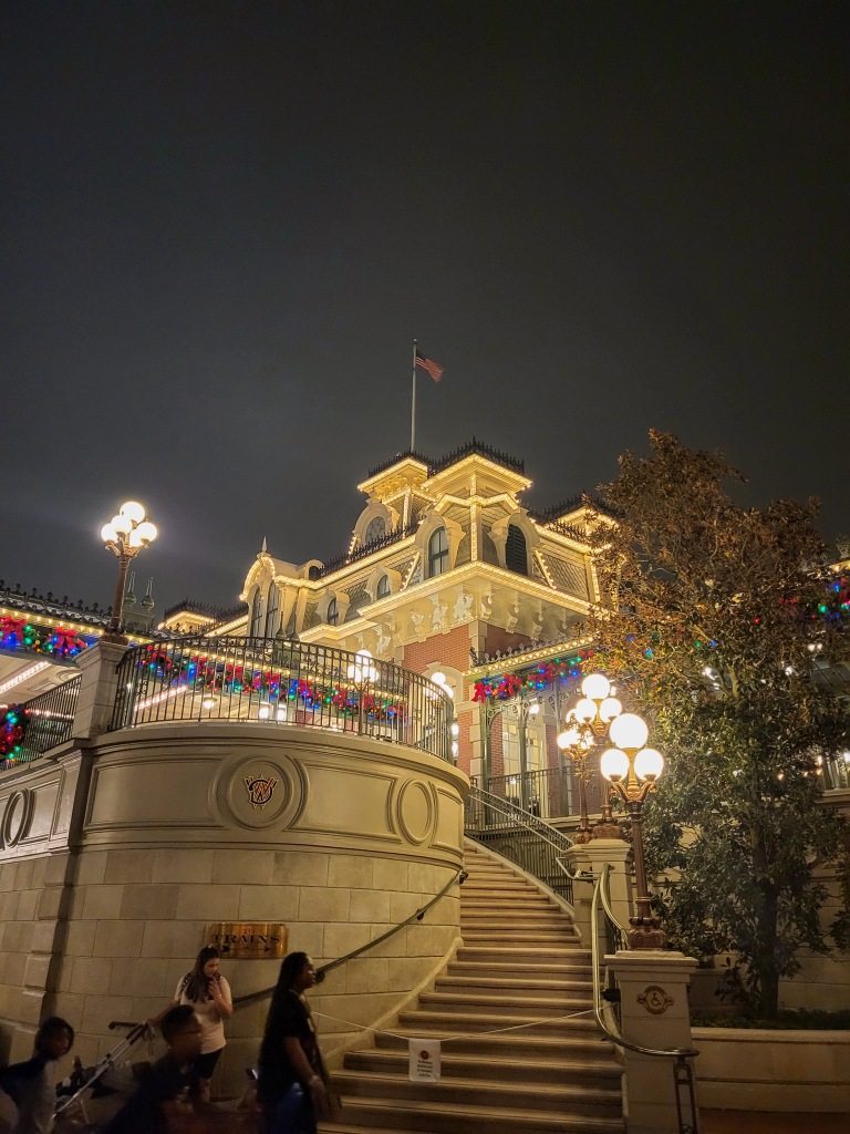 A white building with a small tower lit up with yellow fairy lights. There are green wreaths with colourful baubles adorning it and the sky is black. Steps lead up to the building. There is an American flag flying from the top. 