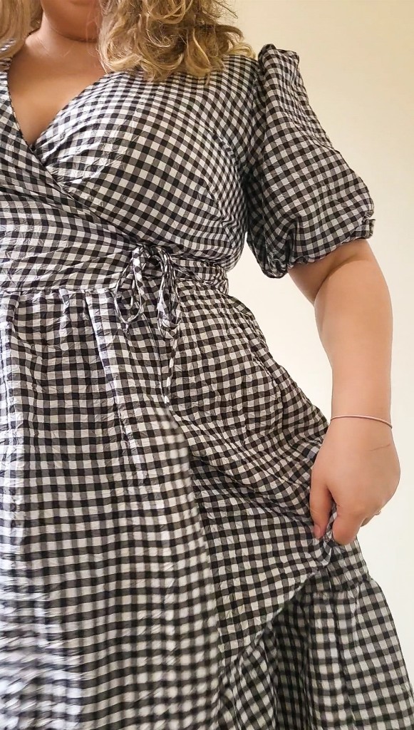 Rosie, a white woman, is wearing a black and white gingham, maxi wrap dress. She is holding the skirt out.