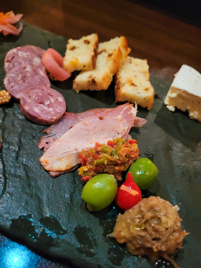 An assortment of pink hams, yellow breads, green olives, and brown sauces on a black slate plate. 