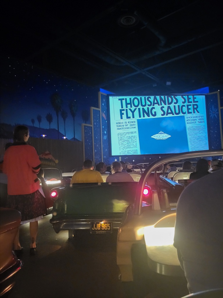 A dark room, where there are the backs of classic cars parked, with people sitting in the cars. There are no roofs to the cars. In front of the cars is a large screen that reads 'thousands see flying saucer'